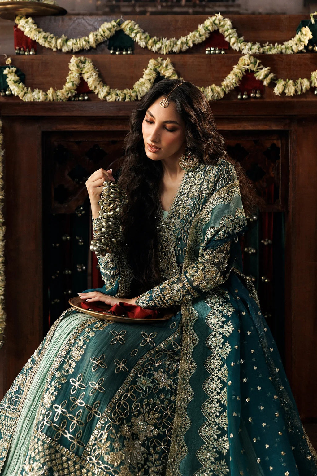 A woman wearing an ornate teal and gold embroidered traditional dress sits in front of a dark wooden fireplace decorated with garlands, holding a tray with red items.