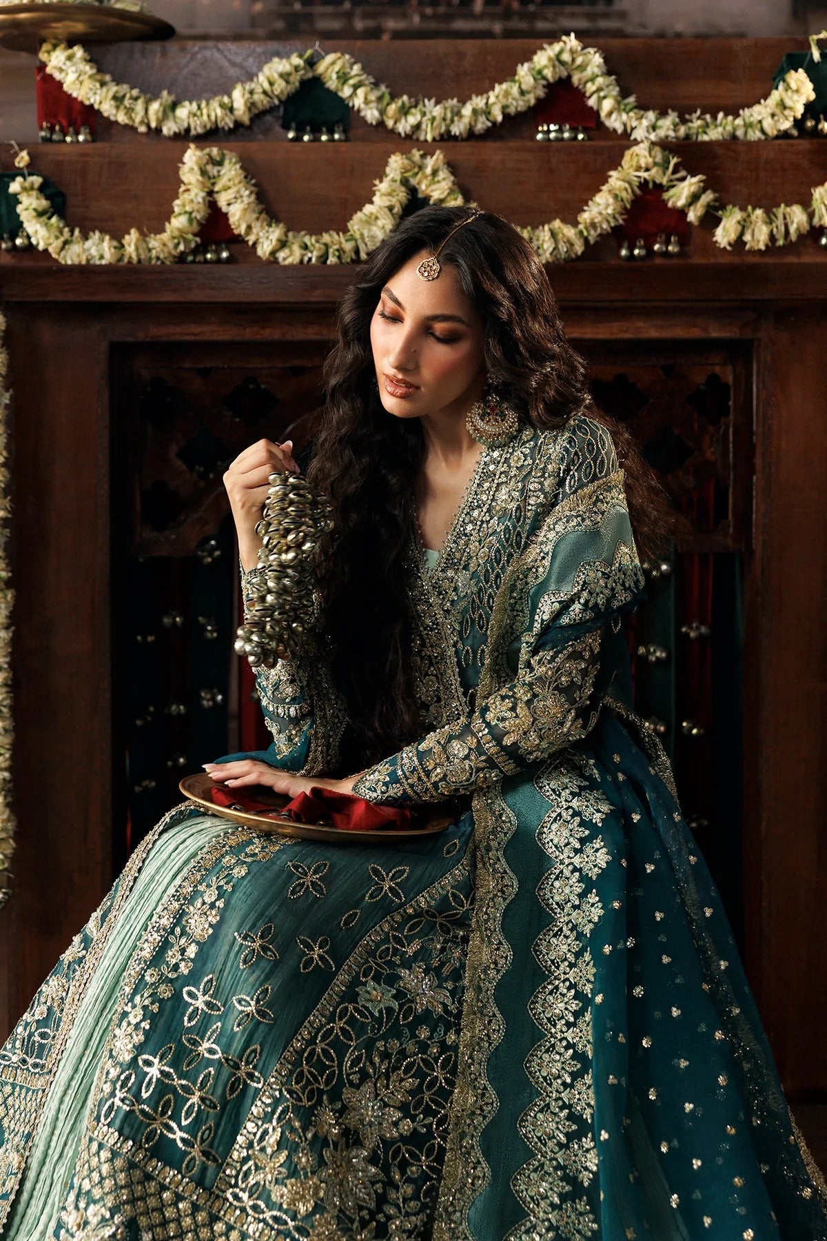 A woman wearing an ornate teal and gold embroidered traditional dress sits in front of a dark wooden fireplace decorated with garlands, holding a tray with red items.