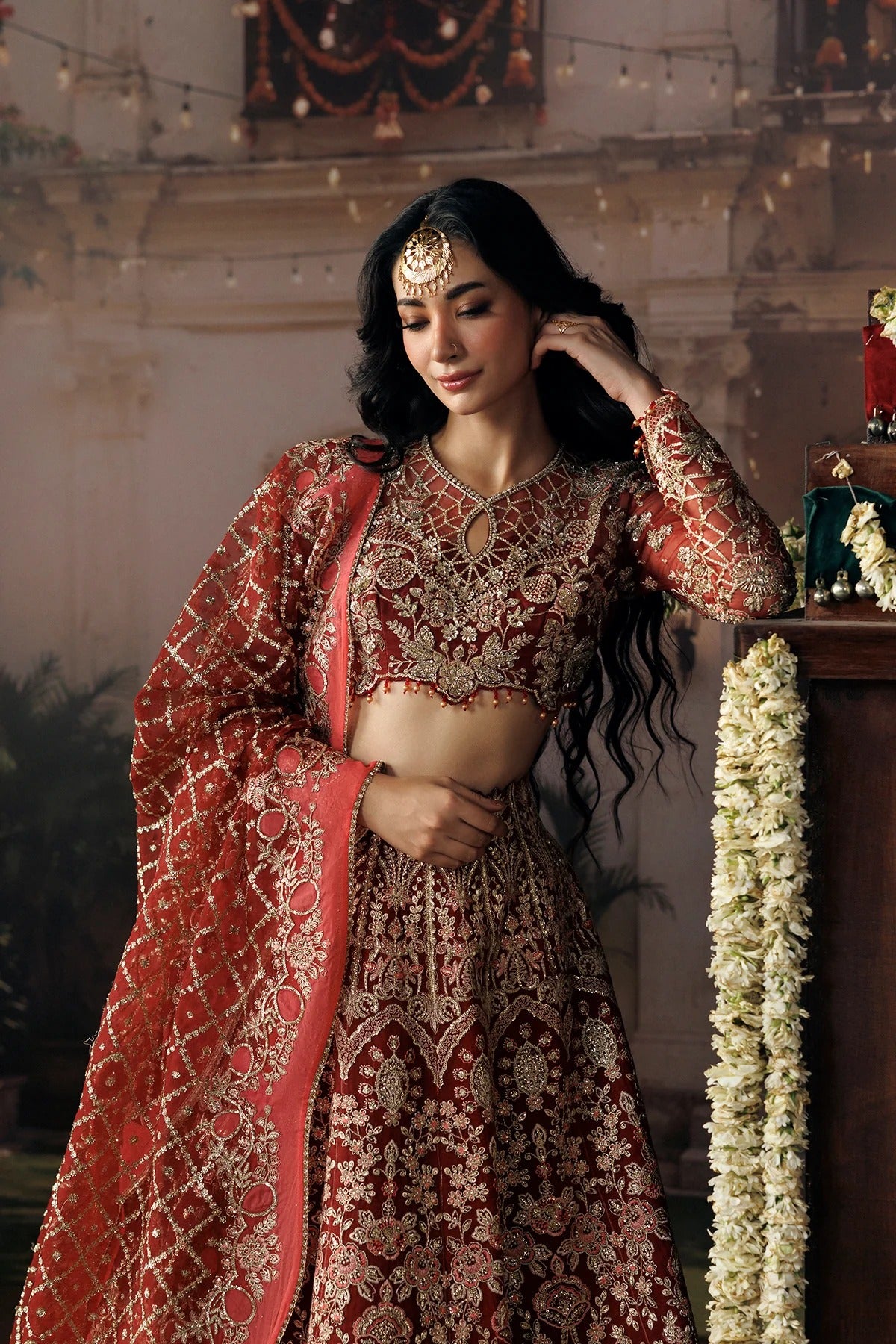 A woman wearing an ornate red and gold embroidered traditional Indian bridal outfit with a matching dupatta, standing indoors against a softly lit decorative background with floral garlands.