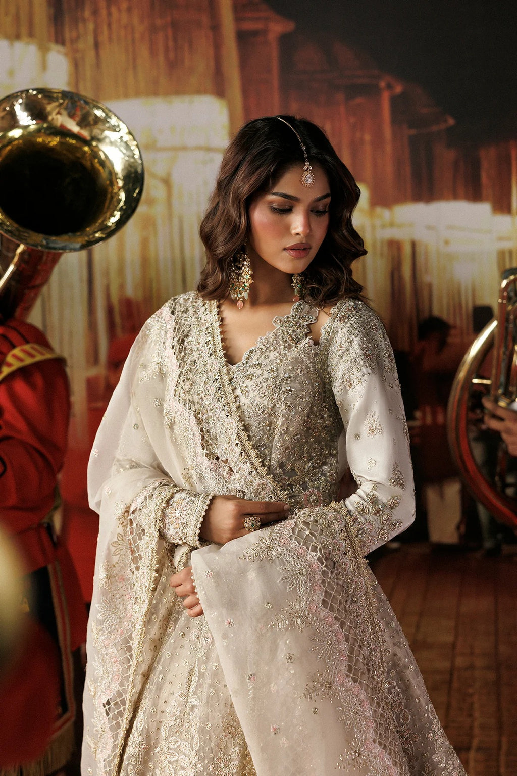 A woman wearing an ornate cream and silver embellished traditional outfit with a matching sheer dupatta, posed in a warmly lit indoor setting with brass instruments partially visible in the background.