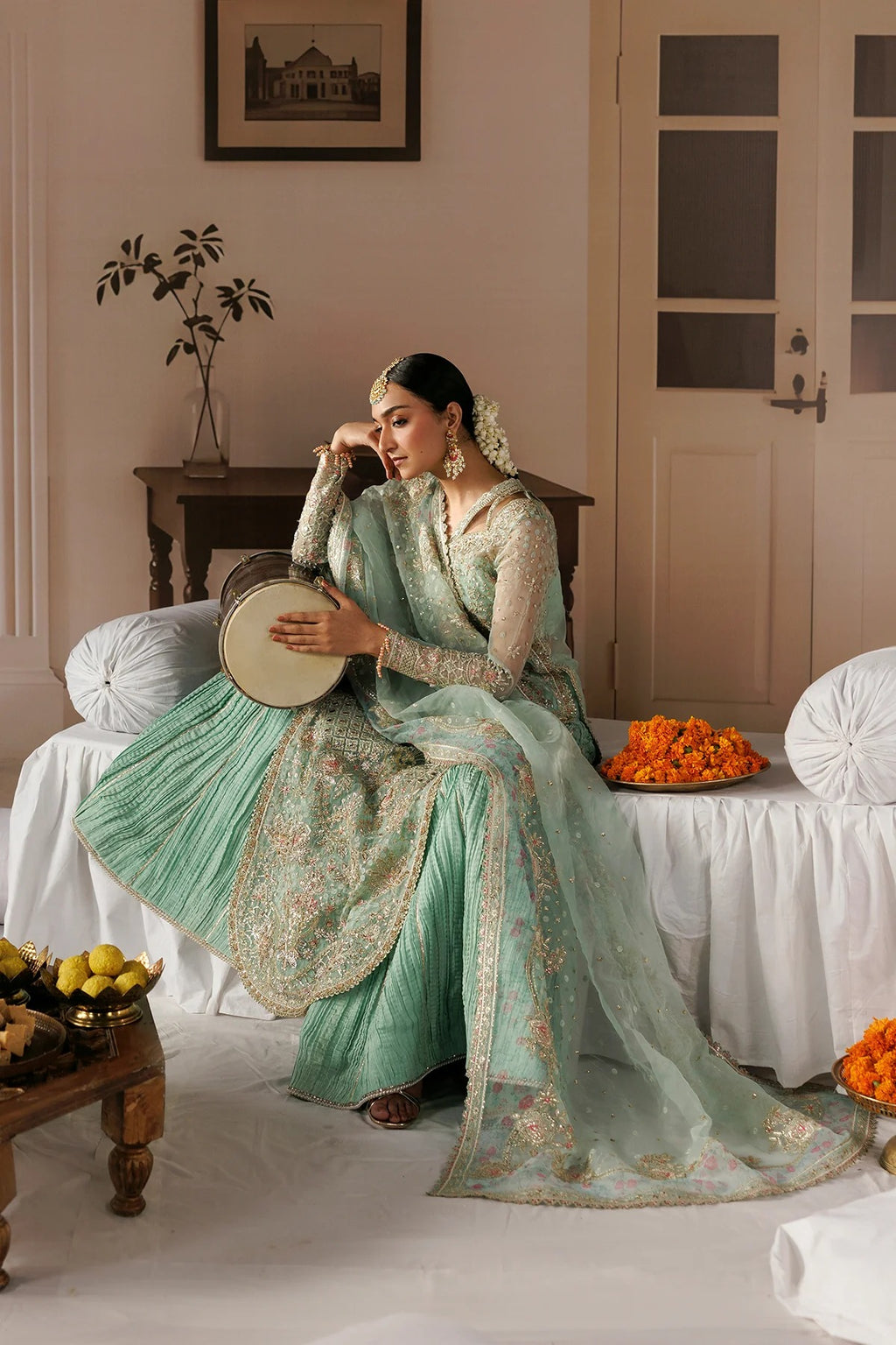 A woman in an ornate mint green and gold embroidered traditional outfit sits on a draped white sofa in a softly lit room, holding a small drum. Decorative flowers and fruit are placed around her, with a framed picture and door in the background.