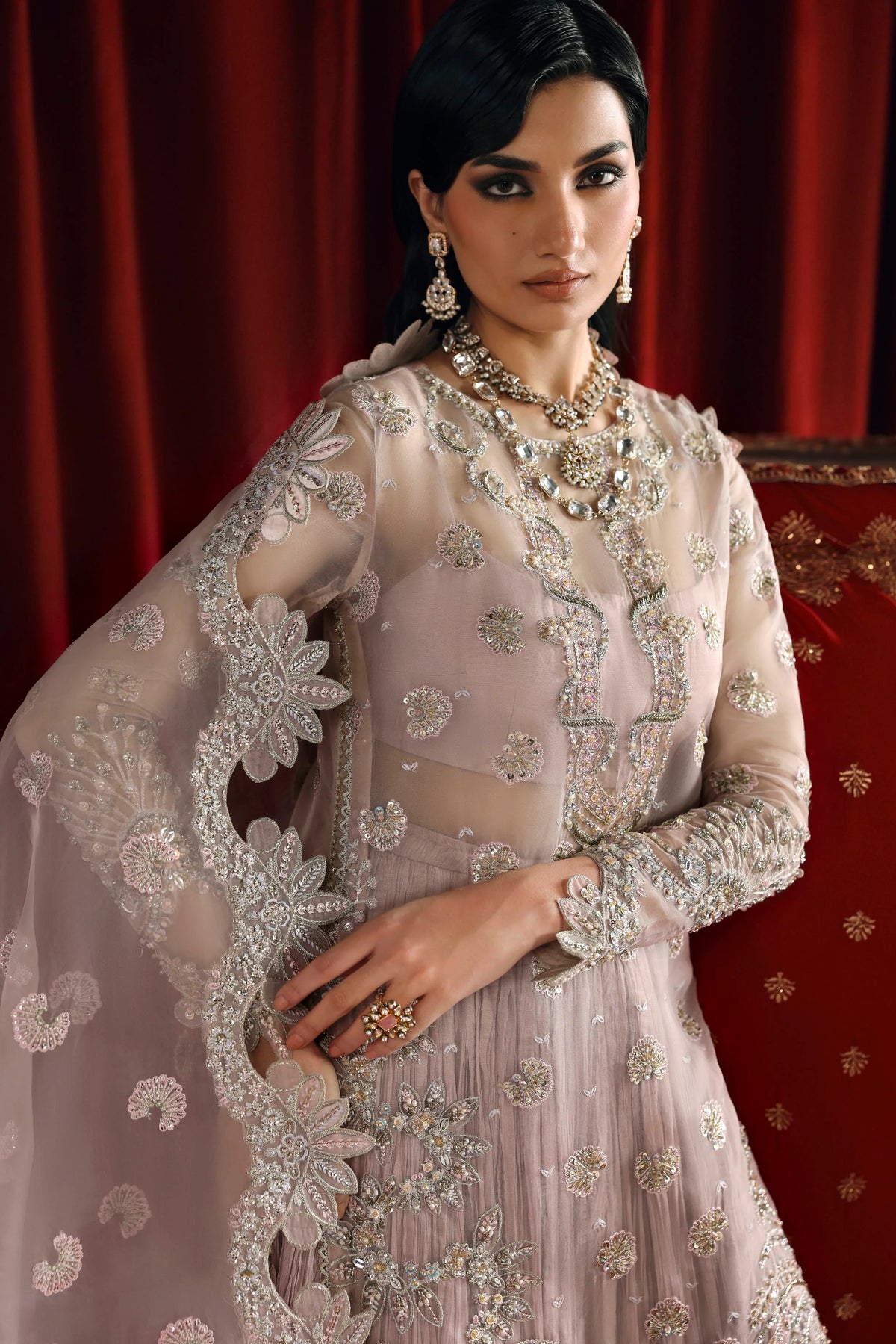 A woman models an ornate light pink traditional outfit with heavy silver and gold embroidery, embellished with intricate jewelry, set against a dark red velvet background.