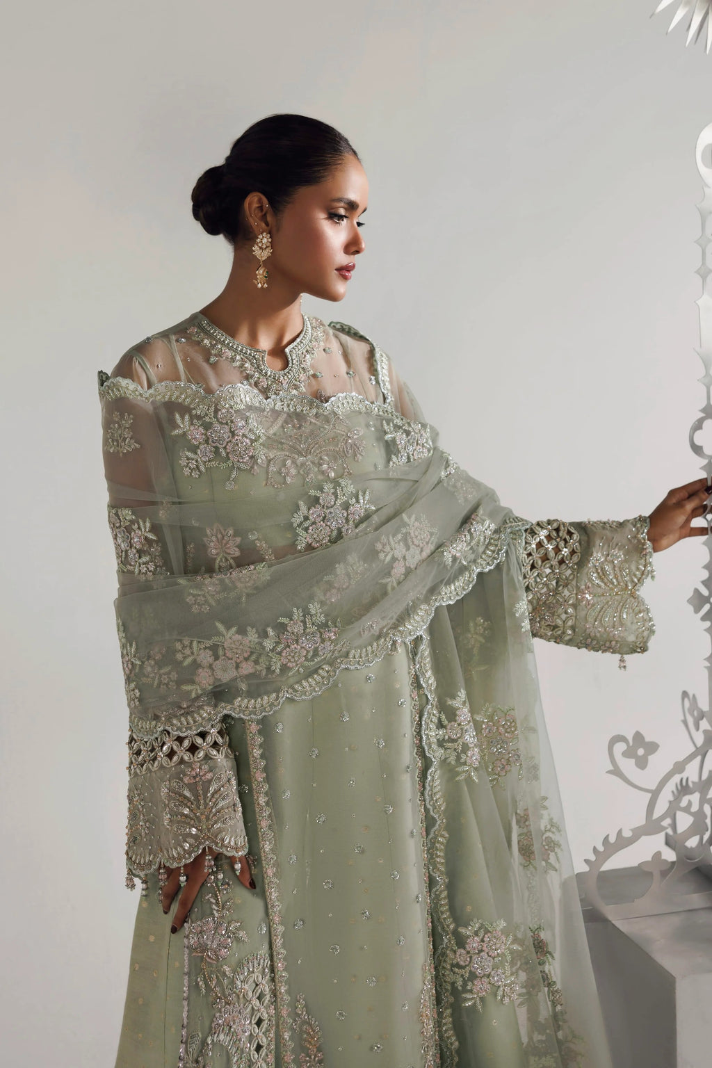 A woman models a traditional light sage green ethnic outfit with intricate floral embroidery and a matching sheer dupatta, posed against a minimalist light gray background.