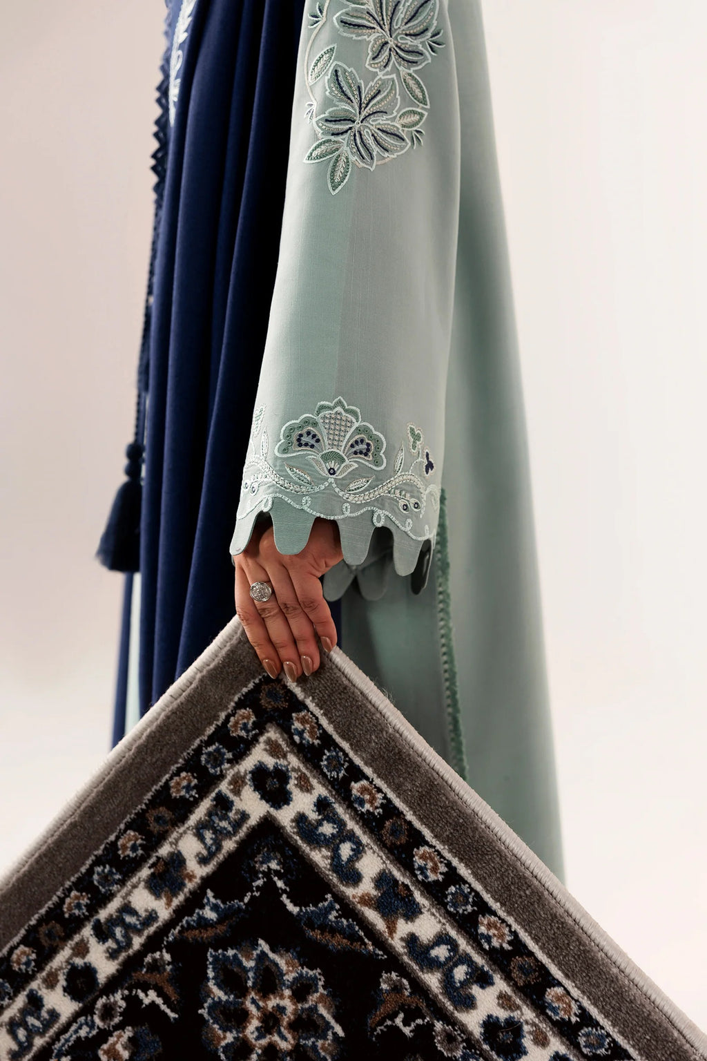 A close-up of a person wearing a light sage green embroidered abaya with scalloped lace cuffs, holding up a dark patterned rug against a plain light background.