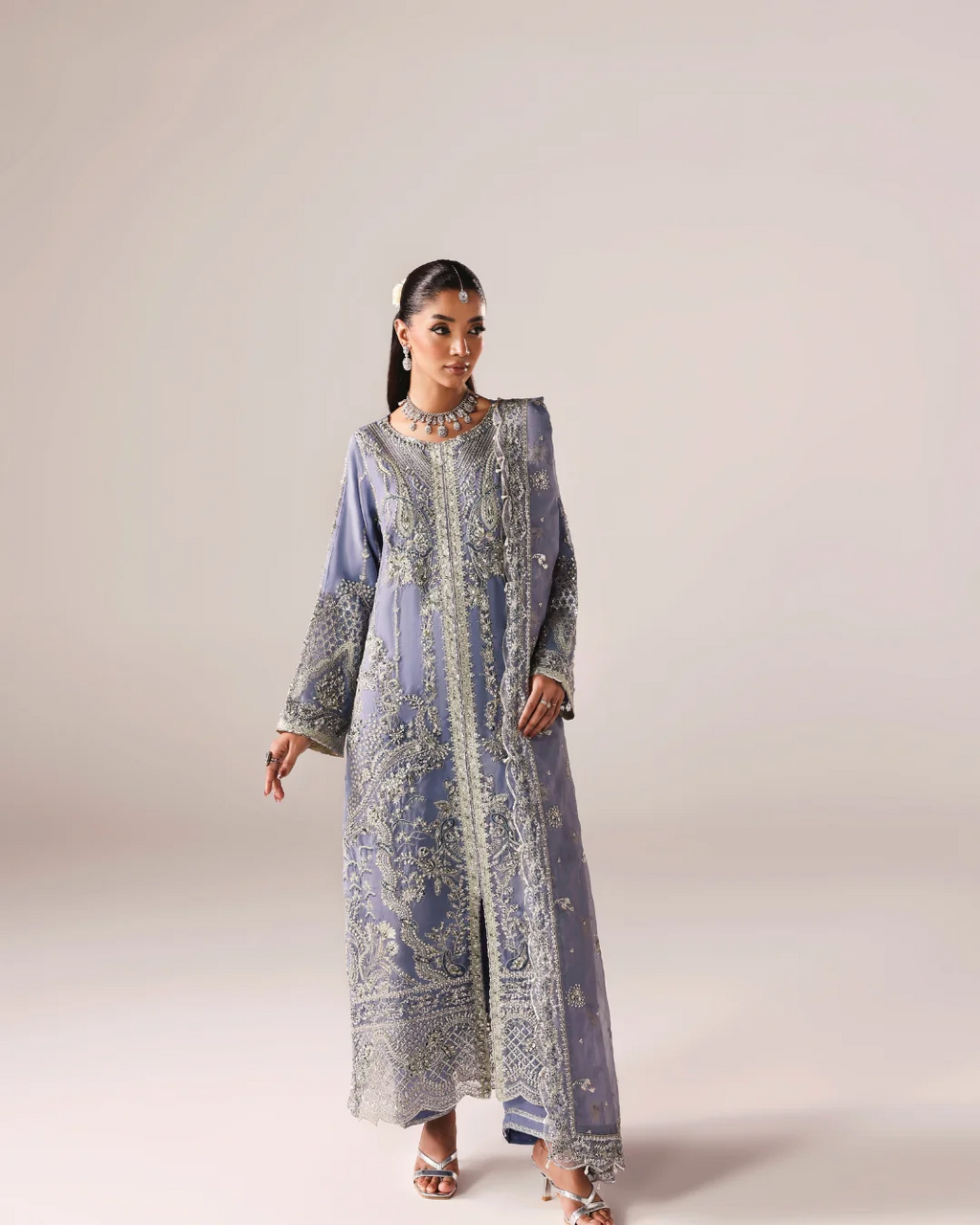 A woman modeling a long, lavender-blue embroidered traditional dress with intricate silver and white detailing, standing against a solid light beige studio background.