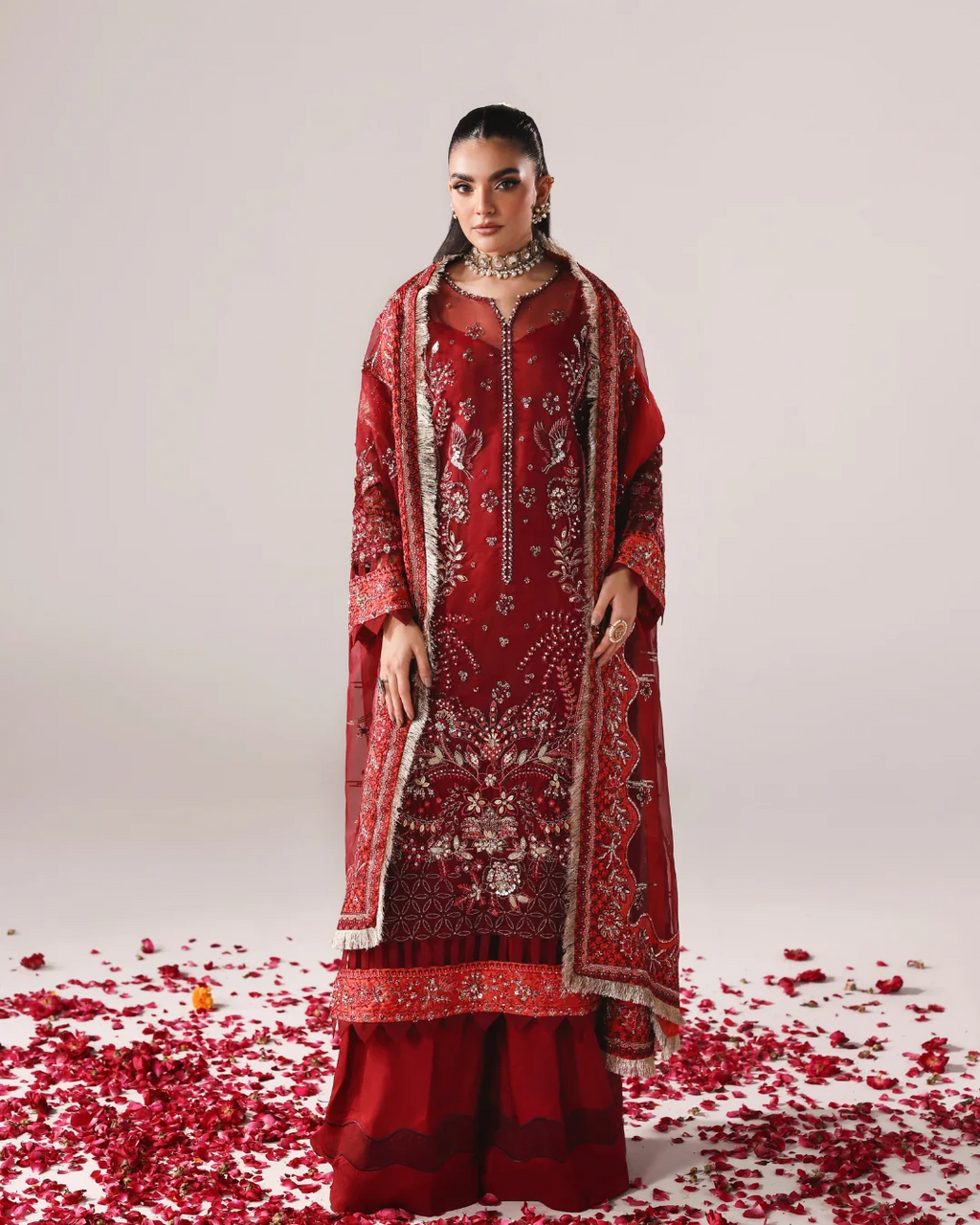 A woman models a traditional red embroidered outfit with a matching sheer dupatta, set against a light gray studio background with scattered red rose petals on the floor.