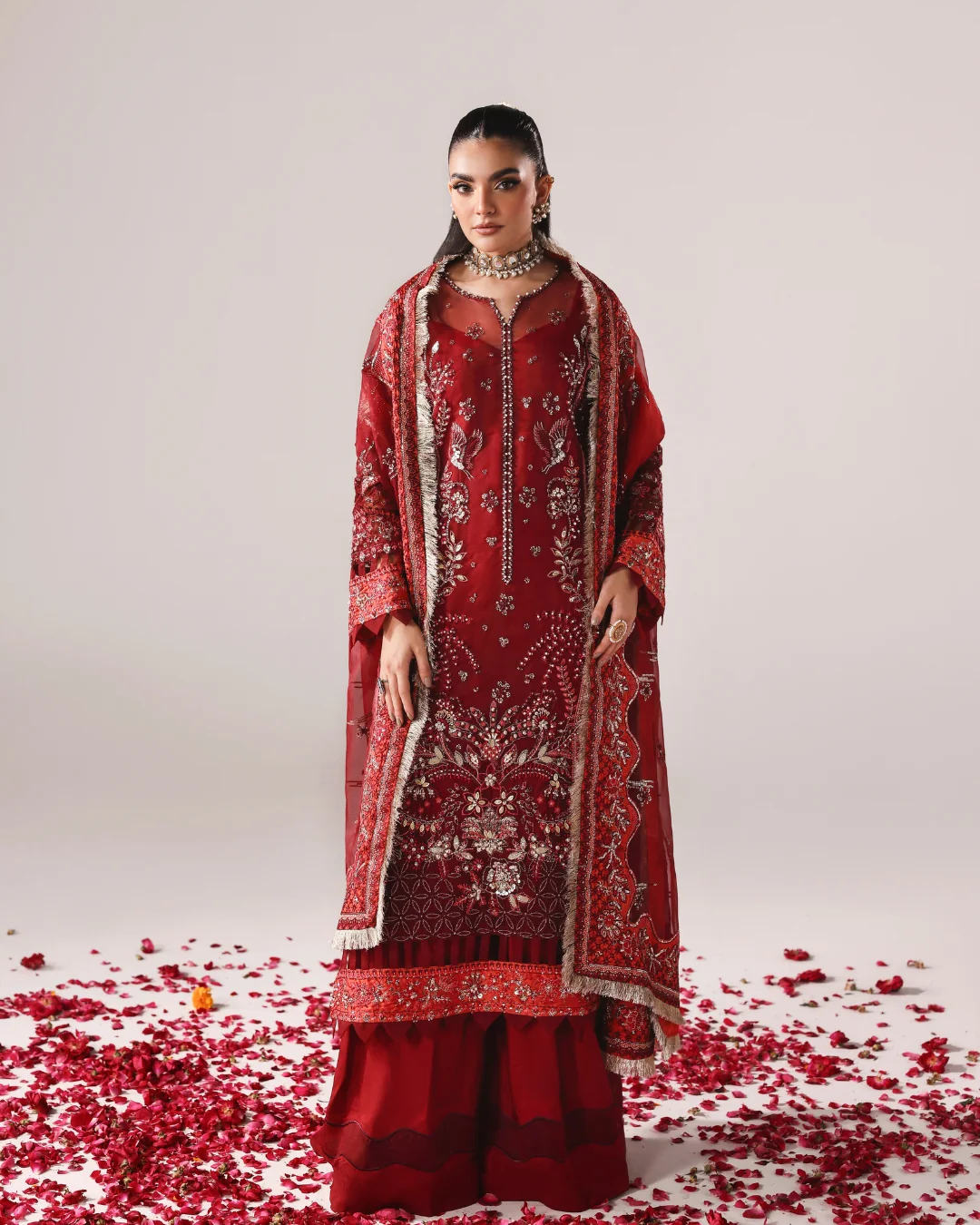 A woman models a traditional red embroidered outfit with a matching sheer dupatta, set against a light gray studio background with scattered red rose petals on the floor.