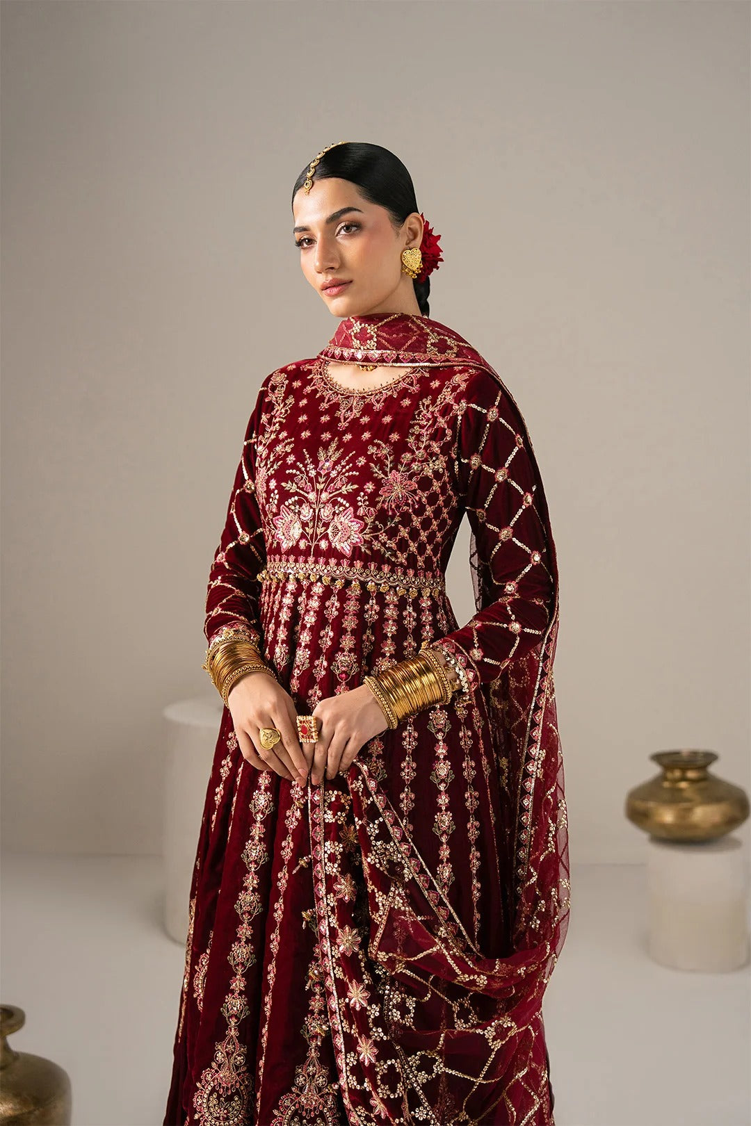 A woman modeling a traditional deep maroon velvet anarkali suit with intricate gold and silver embroidery, styled with gold jewelry against a neutral studio background.