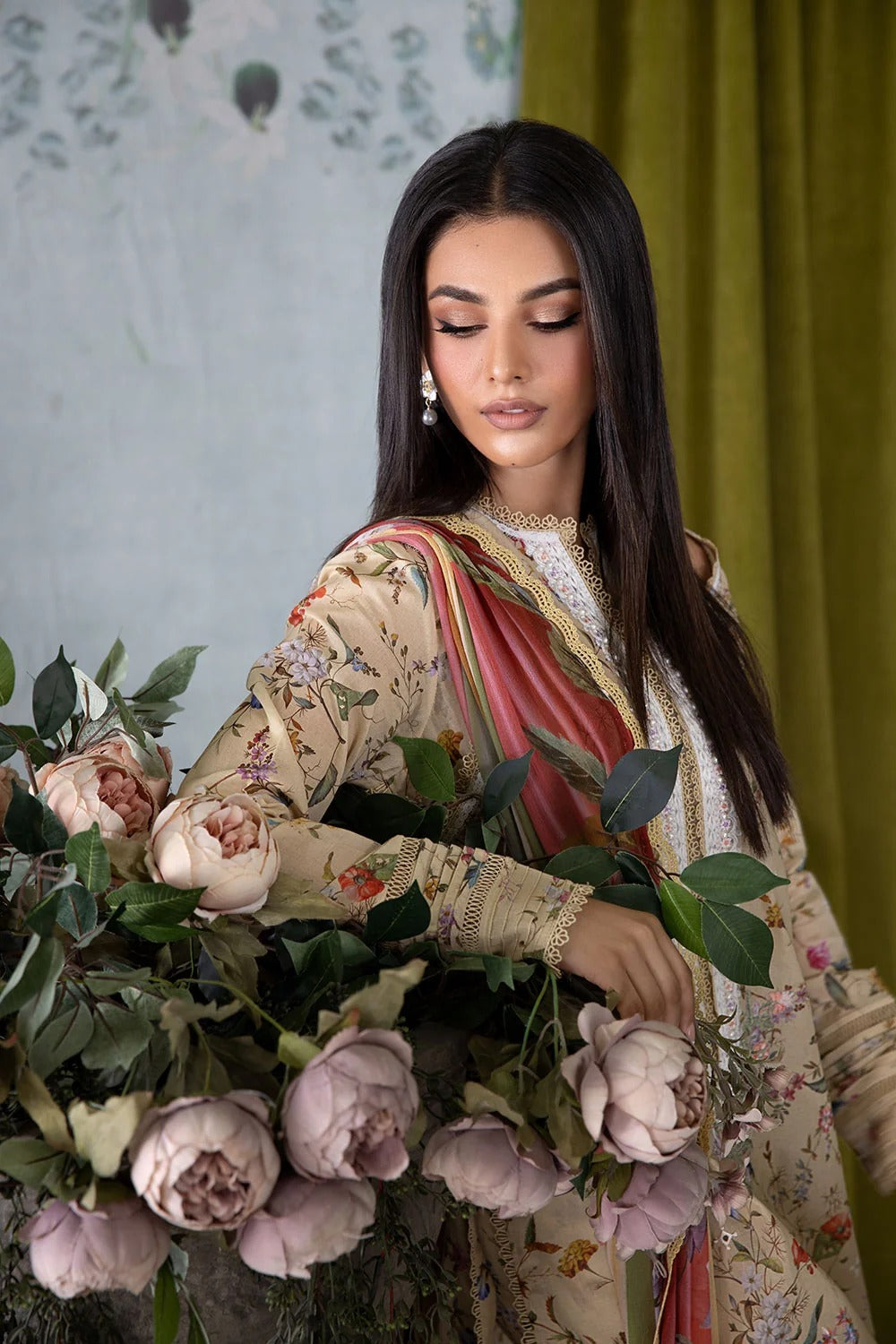 Woman in a floral outfit holding flowers against a green curtain and patterned wall.