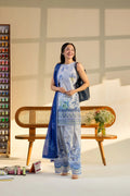 Woman in a blue and white traditional outfit standing in a room with a wooden bench and shelves.