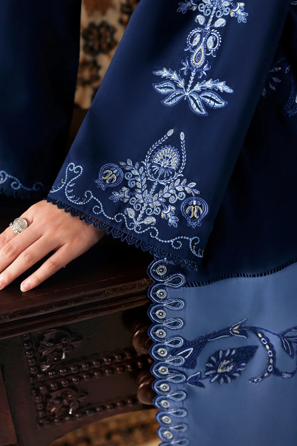 A close-up of a navy blue traditional garment with intricate light blue and white floral embroidery, featuring a scalloped border and a hand resting on a dark wooden surface.