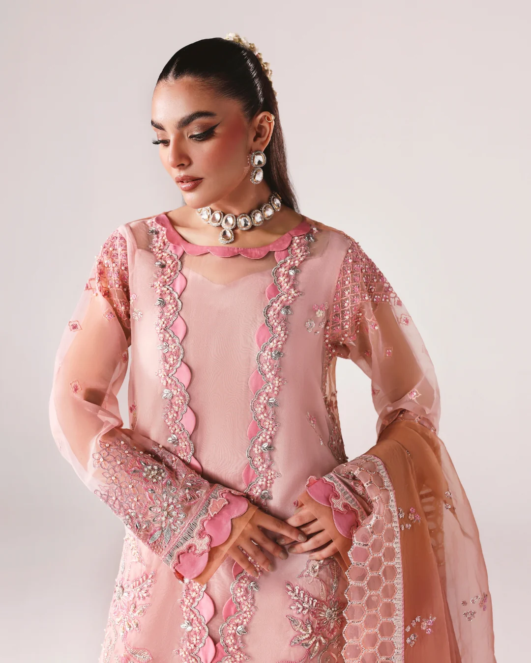 A woman models a light pink traditional embroidered outfit with sheer sleeves, scalloped pink borders, and ornate embellishments, paired with a peach dupatta against a plain light background.