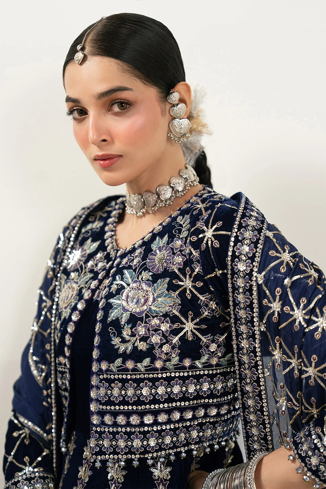 A woman wearing an ornate navy blue embroidered traditional outfit with intricate silver, purple, and white floral detailing, paired with heavy silver jewelry against a plain light background.