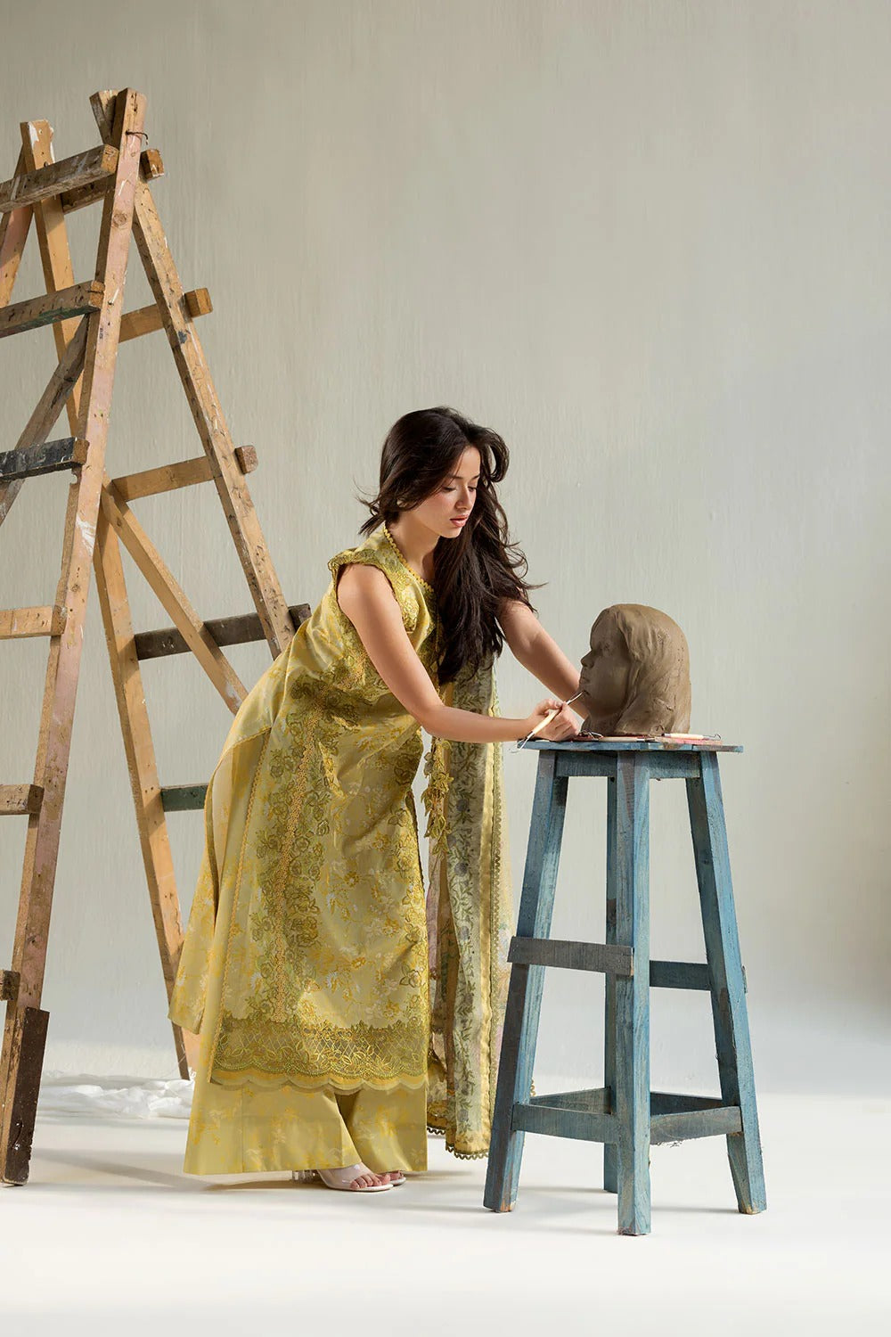 Woman in a yellow dress interacting with a sculpture on a stool against a plain background