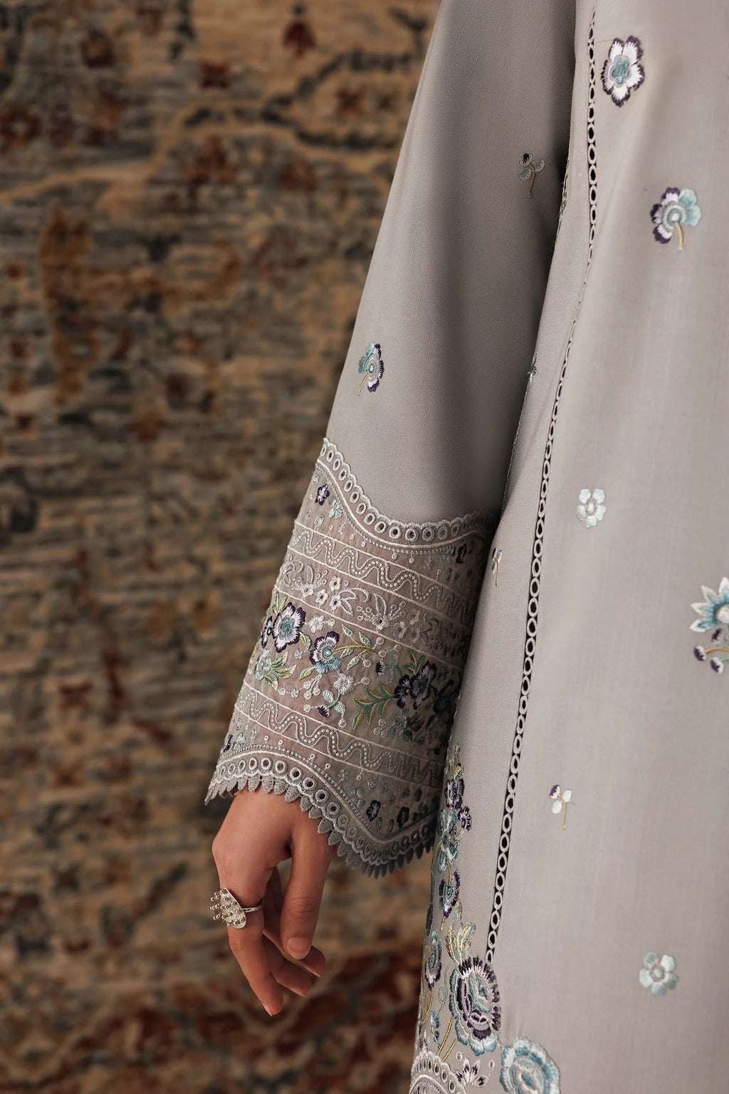 A close-up of a person wearing a light taupe-gray traditional embroidered garment, showing the sleeve with detailed floral embroidery and a ring on their hand, set against a patterned rug.