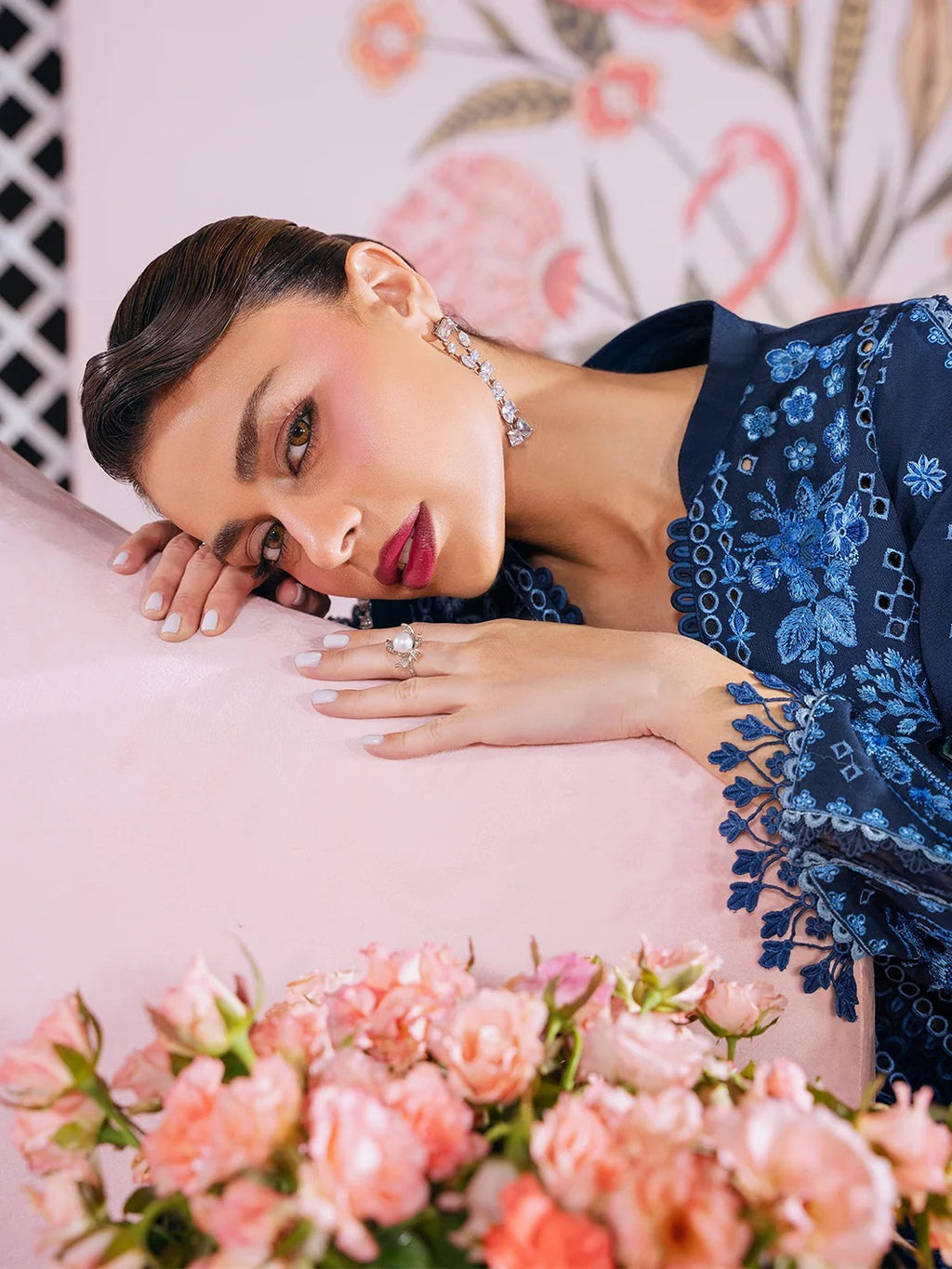 A woman reclining on a pink cushion, wearing a navy blue embroidered traditional outfit with floral patterns and matching jewelry. Pink roses are in the foreground, and a floral-patterned backdrop is visible behind her.