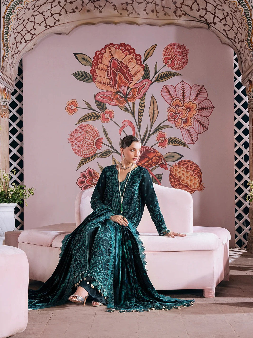 A woman wearing a dark teal embroidered traditional outfit sits on a light pink sofa in front of a decorative floral wall mural.
