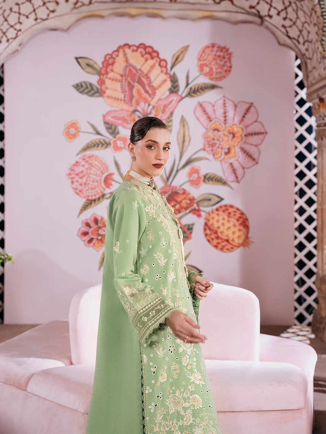 A woman models a light green traditional embroidered outfit with gold detailing, standing in front of a pink floral wall backdrop and a light pink sofa.
