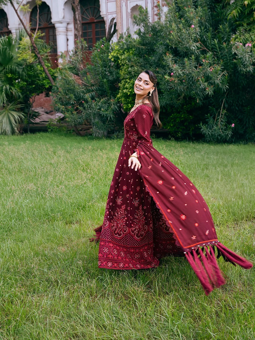 A woman wearing a long maroon embroidered dress stands on a grassy lawn outdoors, with trees and a building visible in the background. The dress has floral embroidery and a flowing skirt with tassel details.