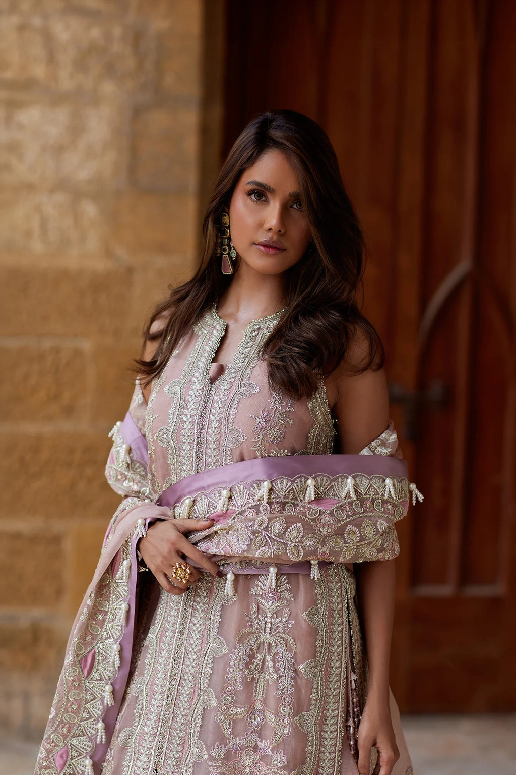 A woman wearing a light pink traditional embroidered outfit with silver mirror work and a lavender sash, standing in front of a stone wall and wooden door.