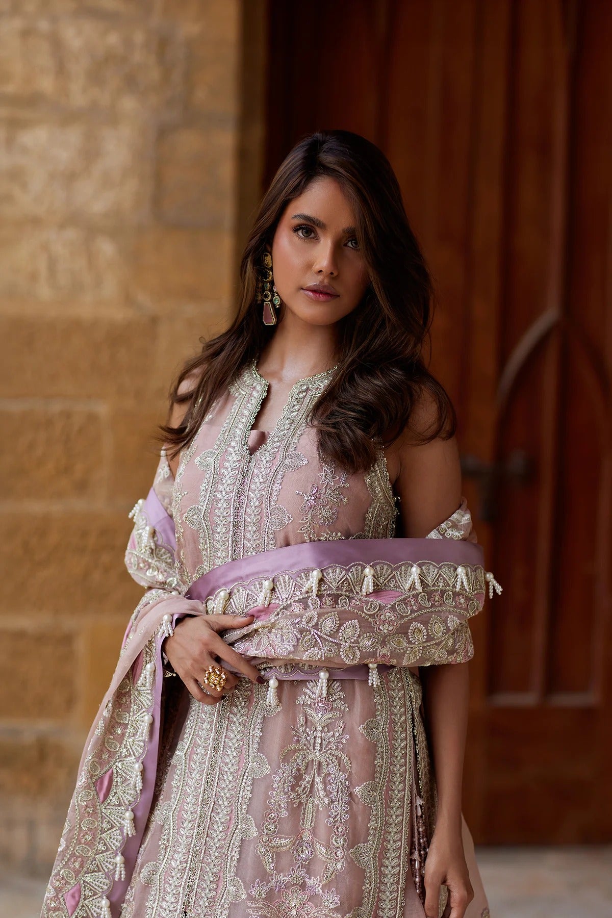 A woman wearing a light pink traditional embroidered outfit with silver mirror work and a lavender sash, standing in front of a stone wall and wooden door.