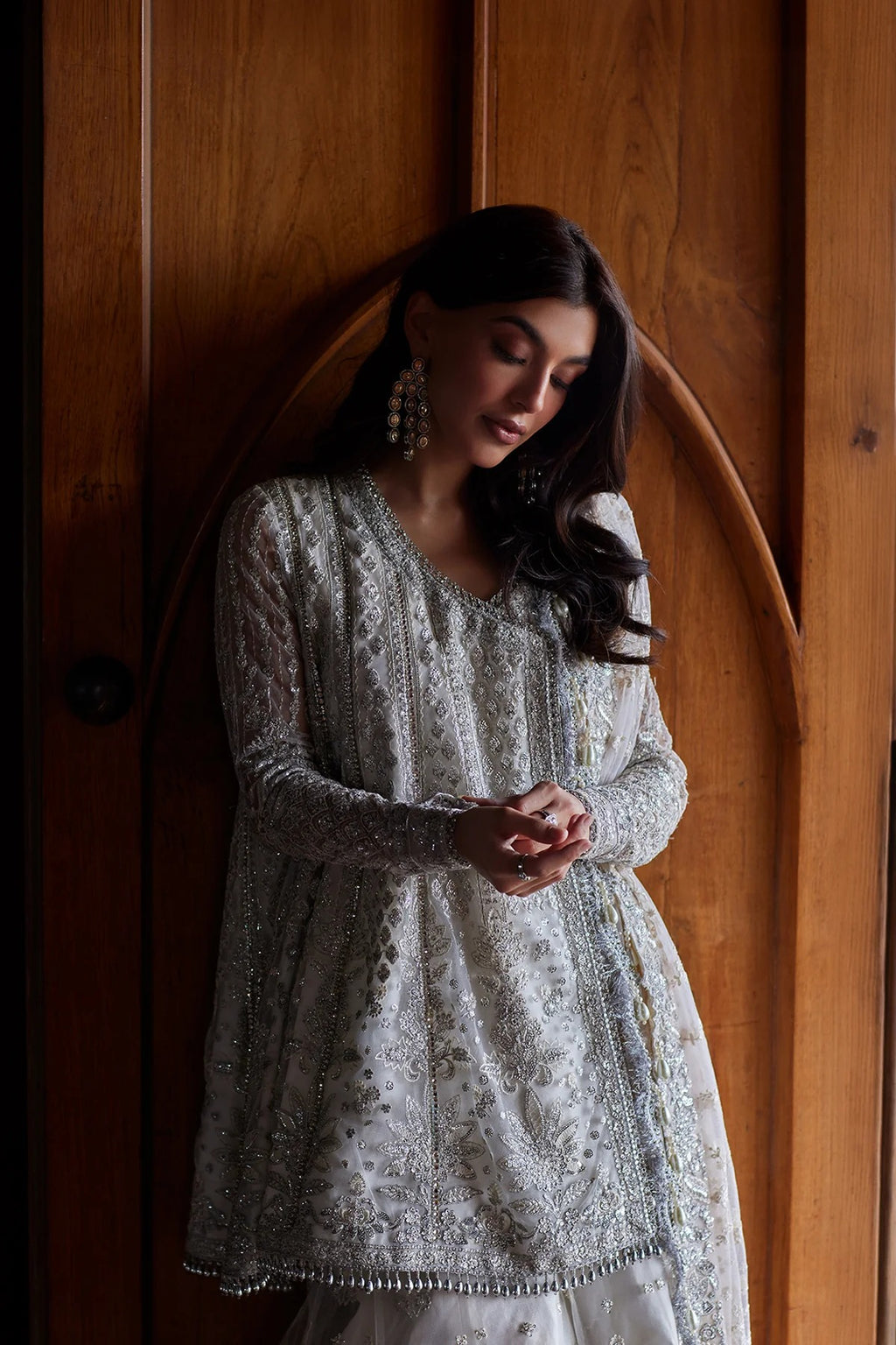 A woman wearing a long-sleeved silver embellished tunic with intricate embroidery, standing in front of a warm wooden door.