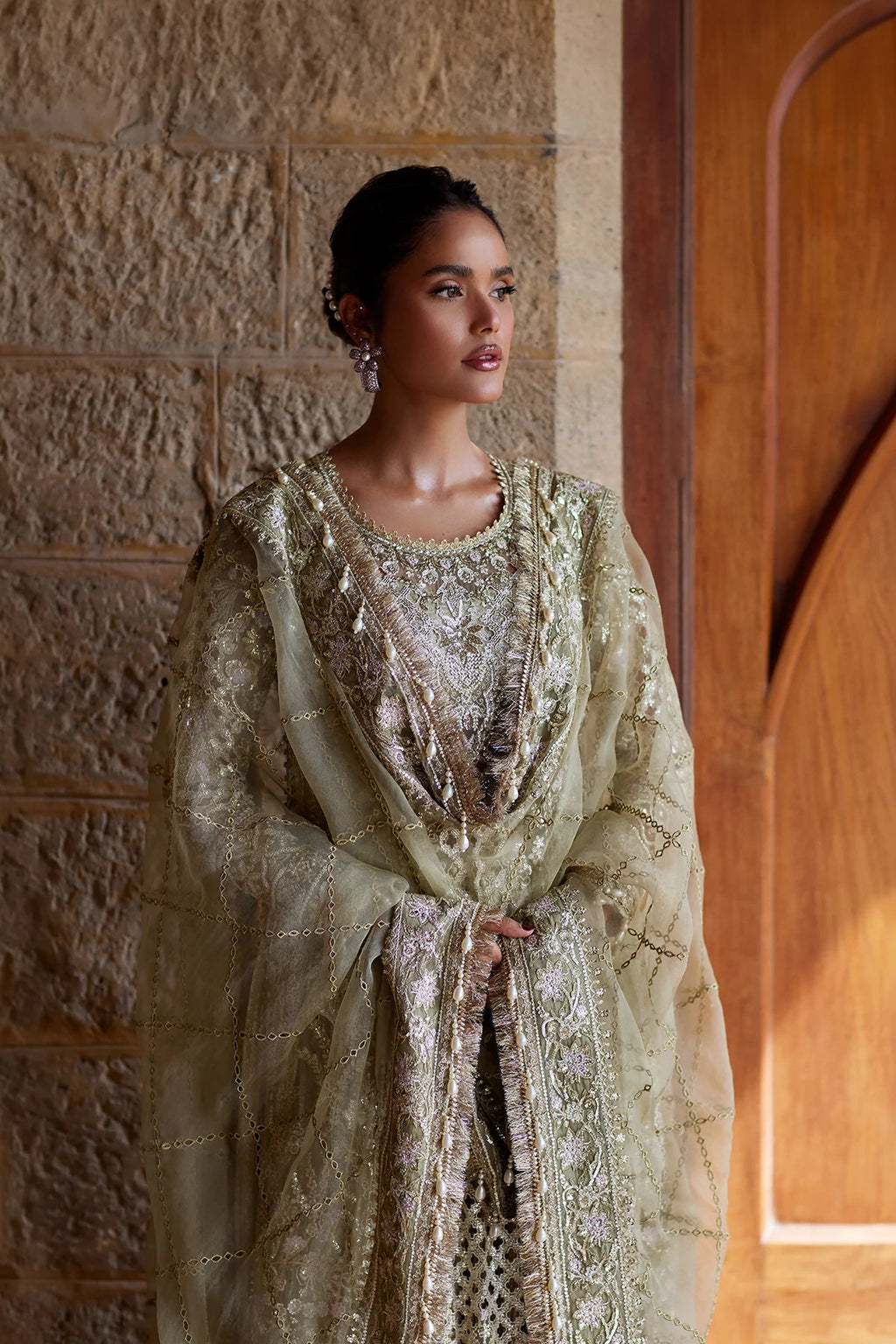 A woman wearing an ornate beige and gold embroidered traditional outfit with a sheer matching shawl, standing against a textured stone wall and wooden door.