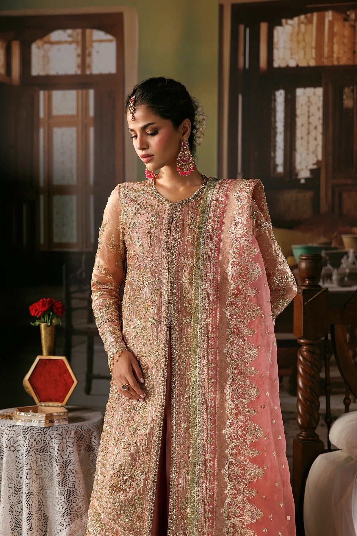 A woman wearing a traditional embroidered peach-pink and beige kurta set with a matching dupatta, standing in an indoor room with wooden furniture and warm ambient lighting.