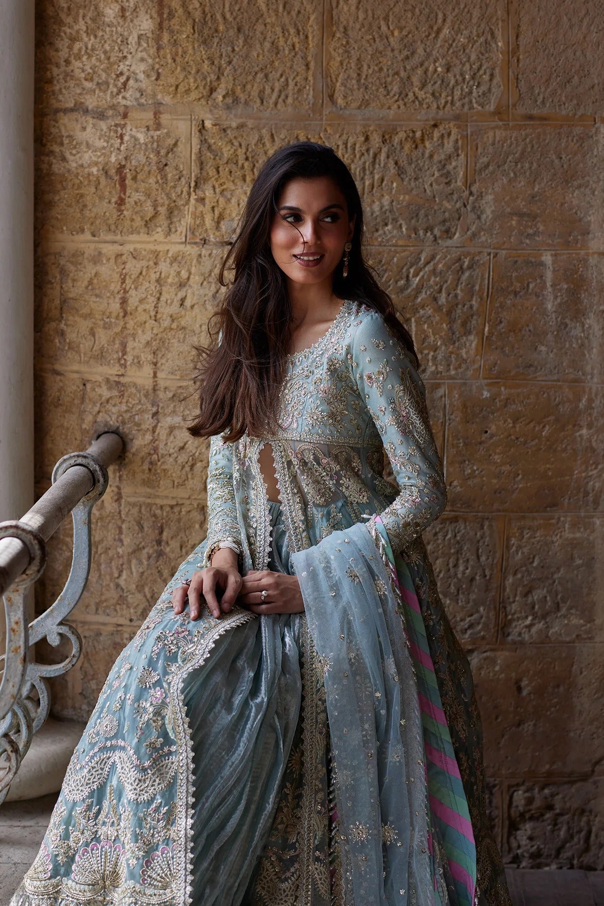 A woman wearing a light blue and silver embroidered traditional outfit with a matching dupatta, posing in front of a textured stone wall.
