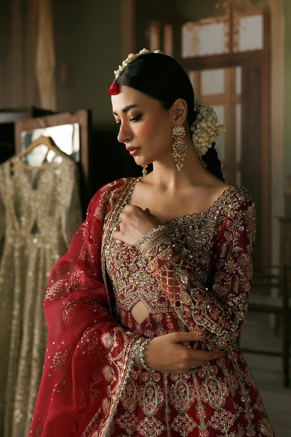 A woman wearing an ornate red and gold embroidered traditional outfit with a matching sheer red dupatta, styled with floral hair accessories and statement earrings in a softly lit indoor setting.