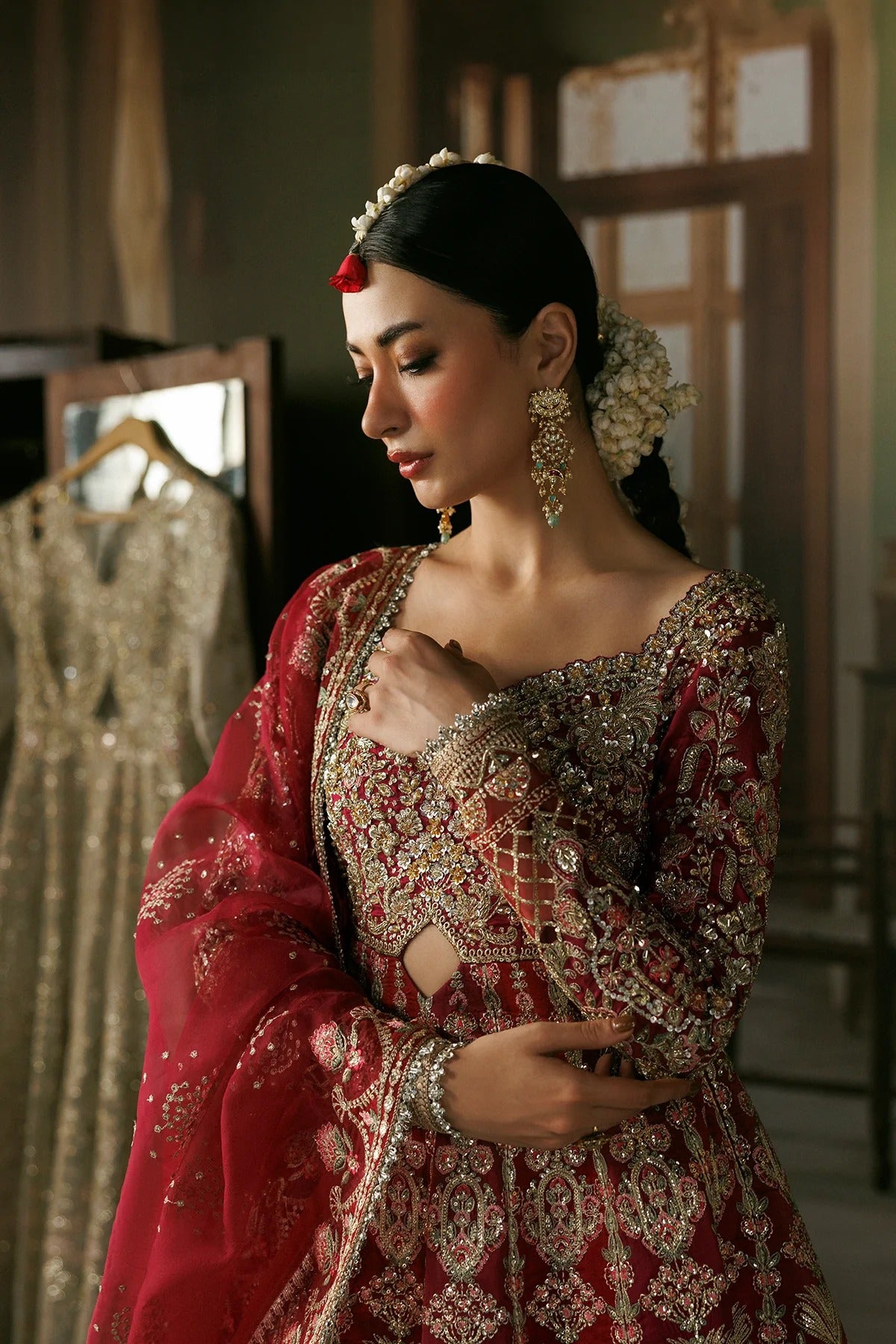 A woman wearing an ornate red and gold embroidered traditional outfit with a matching sheer red dupatta, styled with floral hair accessories and statement earrings in a softly lit indoor setting.