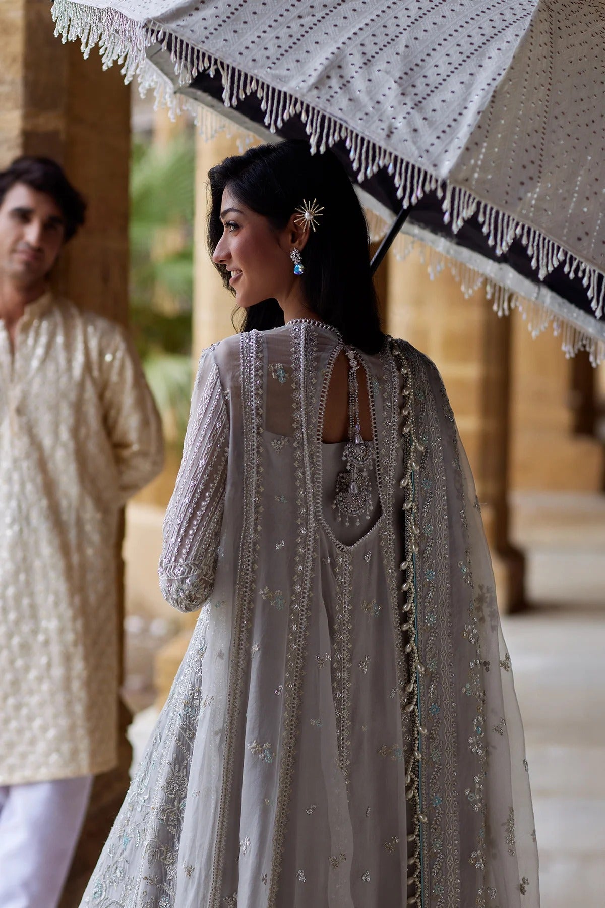 A woman wearing a heavily embroidered light beige or silver traditional outfit stands under a white fringed parasol, shown from the back. A man in a cream kurta is blurred in the background near architectural columns.