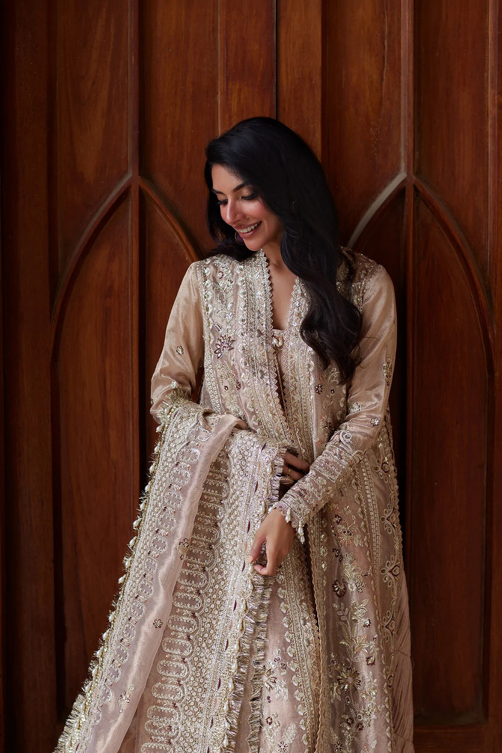 A woman wearing a beige and gold heavily embroidered traditional outfit with a matching dupatta, standing in front of a dark wood paneled background.