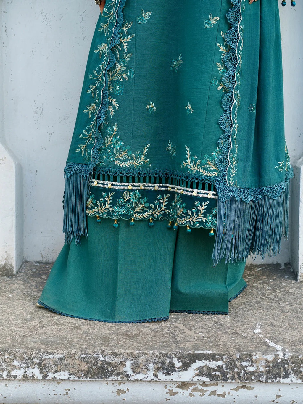 A close-up of a teal green traditional outfit with matching dupatta, featuring intricate gold and silver floral embroidery, lace borders, and tassels, draped over pleated trousers against a rustic concrete wall.