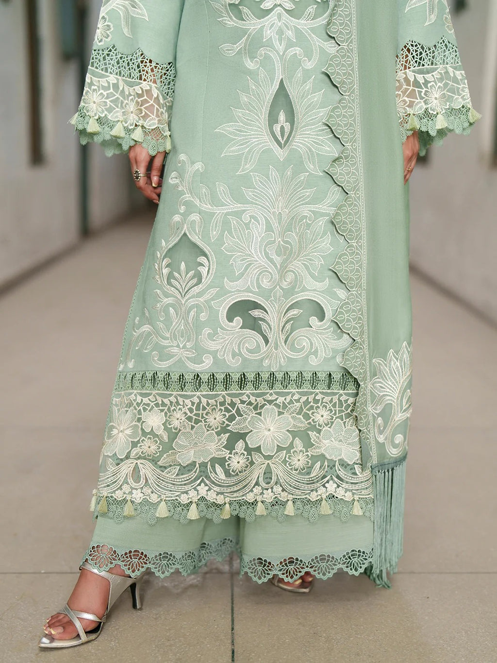 A person wearing a mint green traditional embroidered outfit with white floral embroidery, lace borders, and tassel details, paired with metallic silver high-heeled sandals in a hallway.
