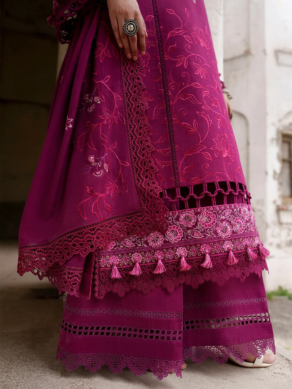 A close-up of a magenta pink embroidered traditional outfit with intricate lace borders and tassels, worn by a model whose hand and feet are partially visible.