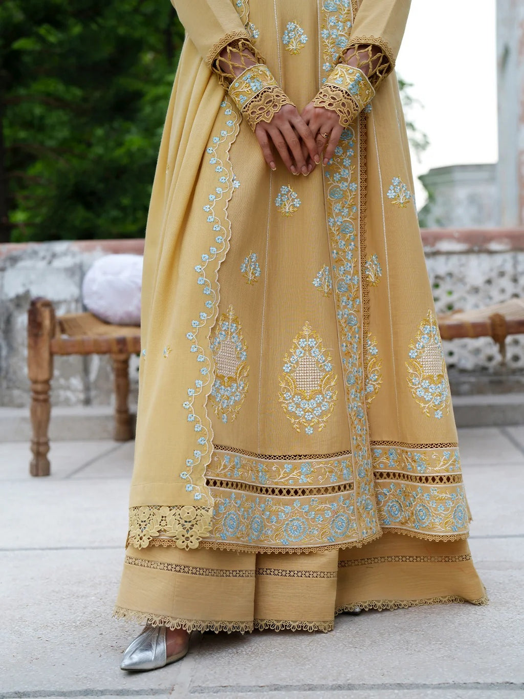 A person wearing a long yellow embroidered traditional dress with intricate gold and light blue floral patterns, standing on a stone terrace.