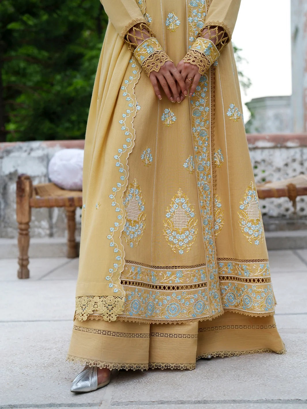 A person wearing a long yellow embroidered traditional dress with intricate gold and light blue floral patterns, standing on a stone terrace.