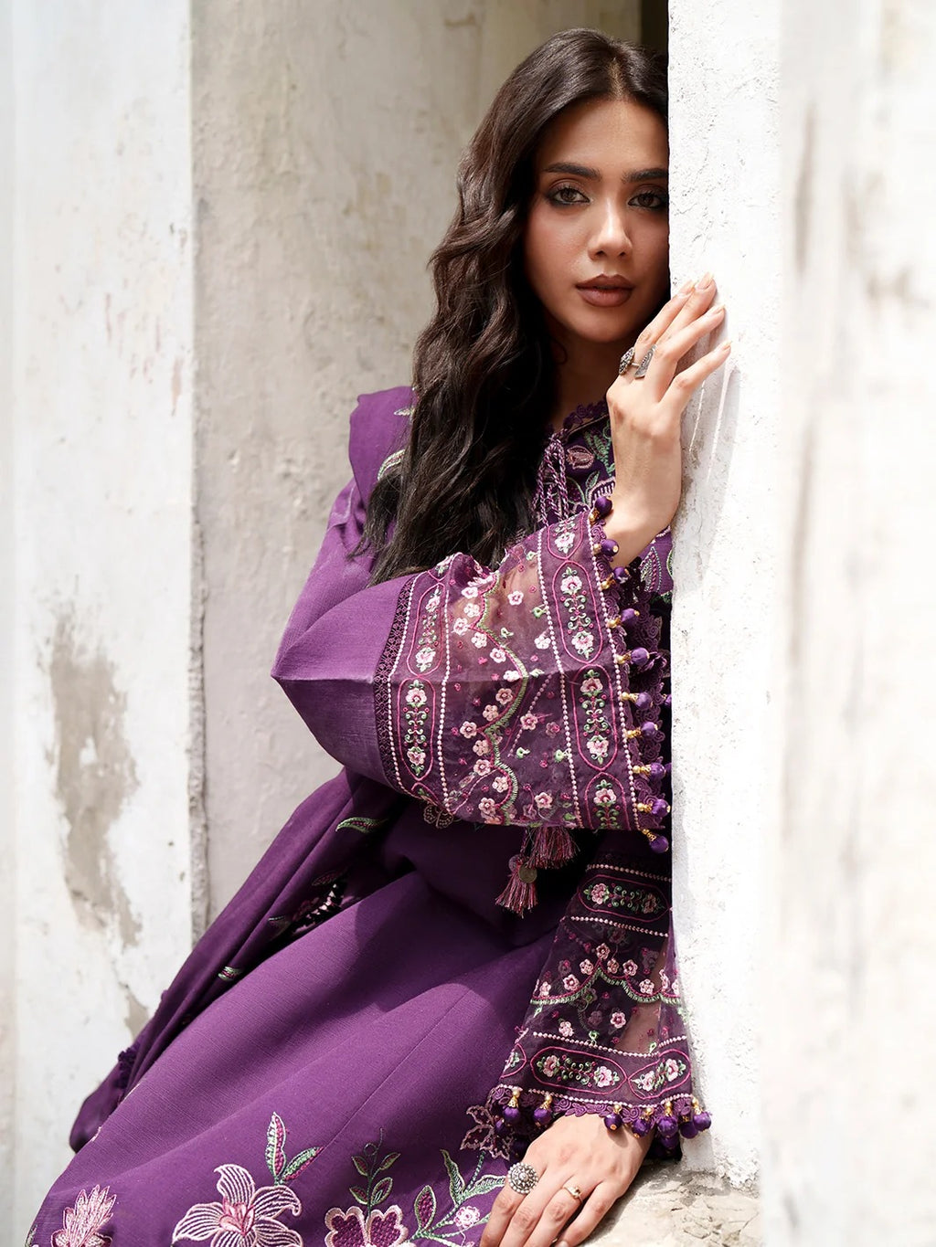 A woman wearing a purple embroidered traditional outfit leans against a textured white wall, with her hand resting on the wall and her hair styled in loose waves.