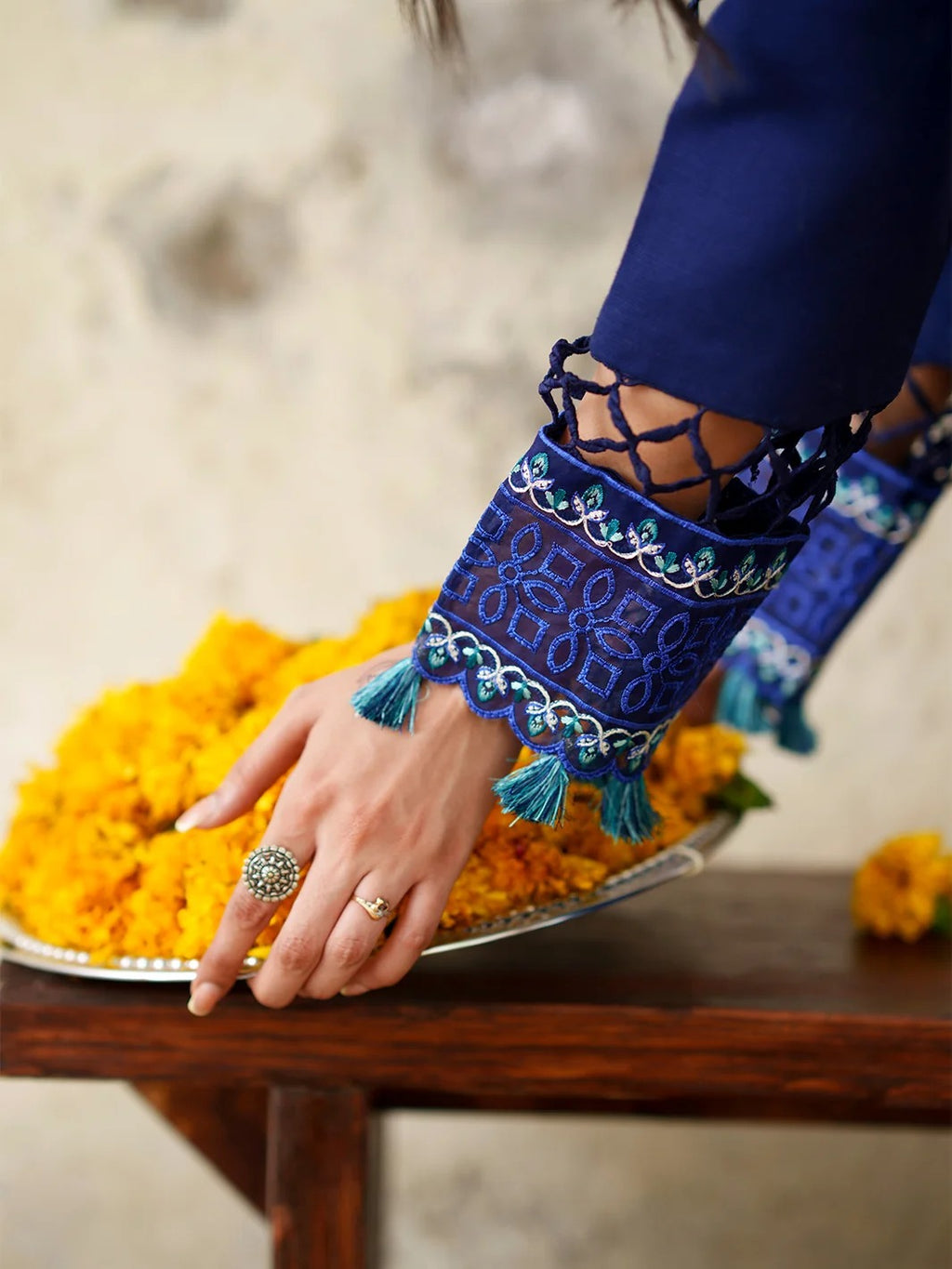 A close-up of a person's hand resting on a silver tray filled with bright yellow marigold flowers, wearing a navy blue embroidered sleeve with lace-up cutouts and tassel details.