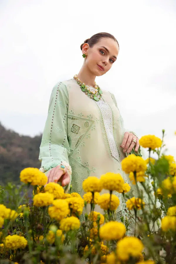 A woman wearing a light green embroidered traditional outfit stands in a field of yellow flowers, looking towards the camera.
