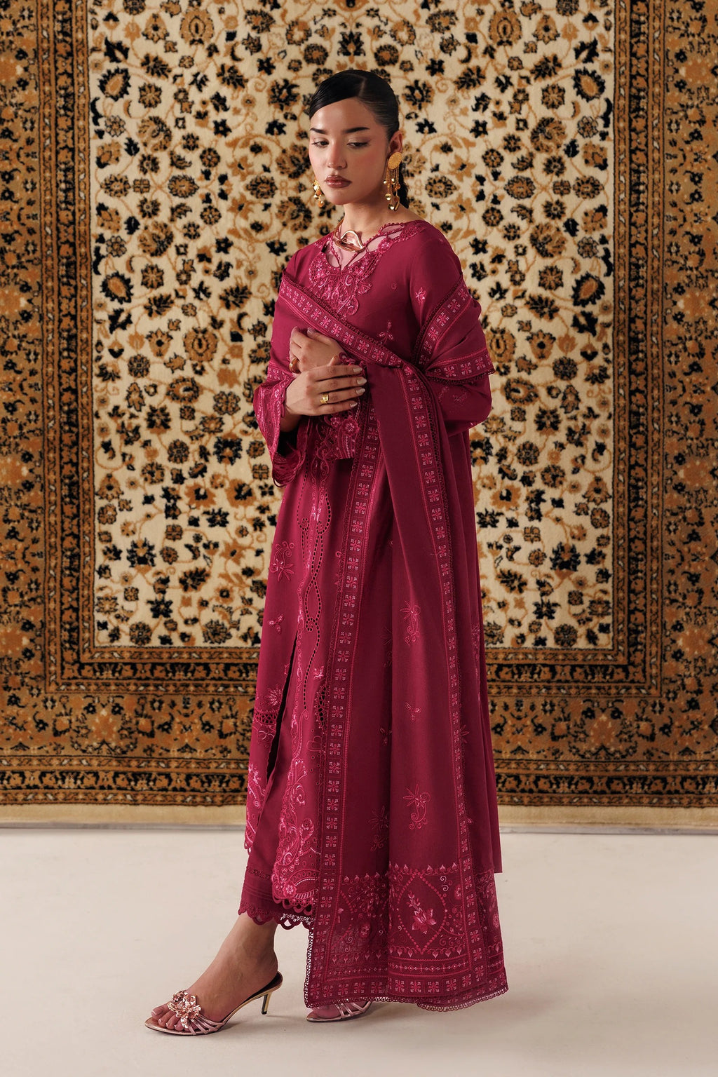 A woman models a traditional magenta pink embroidered kurta set with a matching dupatta, standing in front of a large patterned oriental rug.