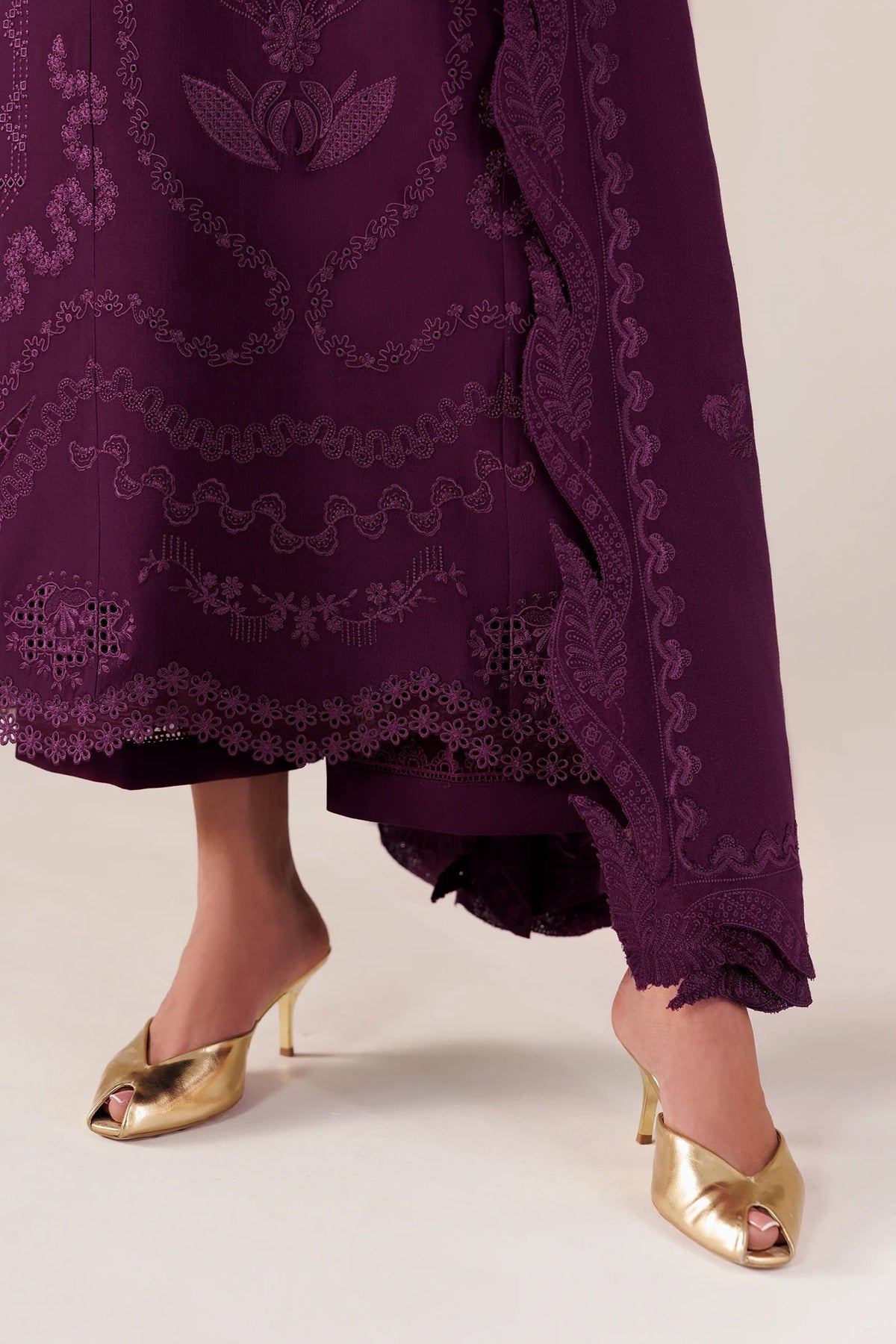 Close-up of a person wearing a deep purple embroidered traditional skirt or drape with matching lace trim and metallic gold mule heels against a plain light beige studio background.