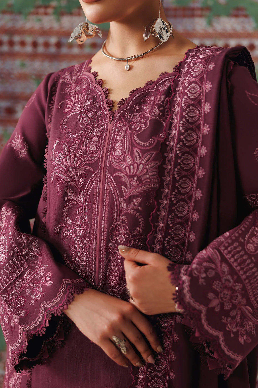 A close-up of a person wearing a deep plum embroidered tunic with lace-trimmed neckline and wide sleeves, styled with statement earrings and a necklace against a patterned backdrop.