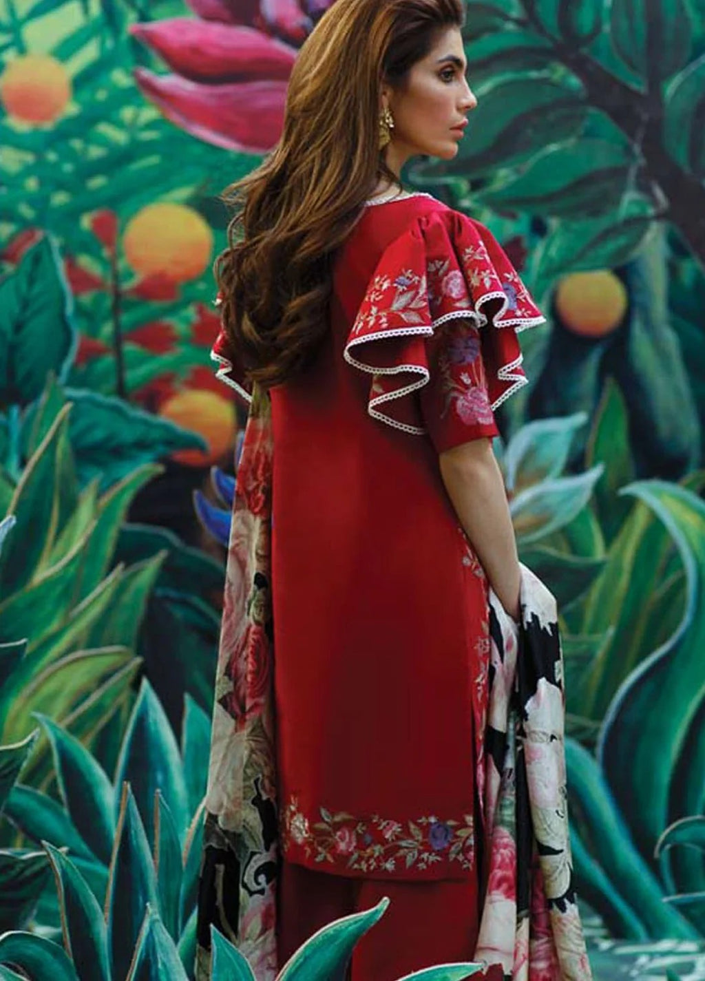 A woman wearing a red traditional outfit with floral-patterned ruffled sleeves and a matching patterned dupatta, posed in front of a colorful tropical garden mural.