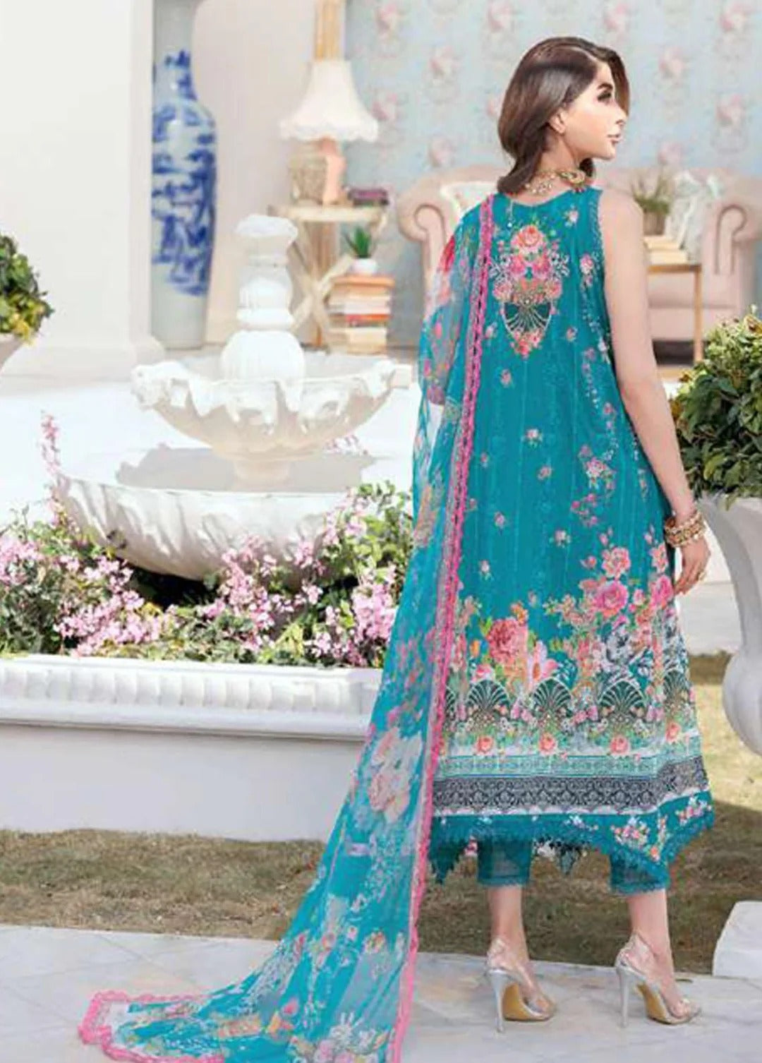 A woman modeling a teal blue ethnic three-piece suit with floral embroidery and a matching dupatta in an outdoor garden setting.