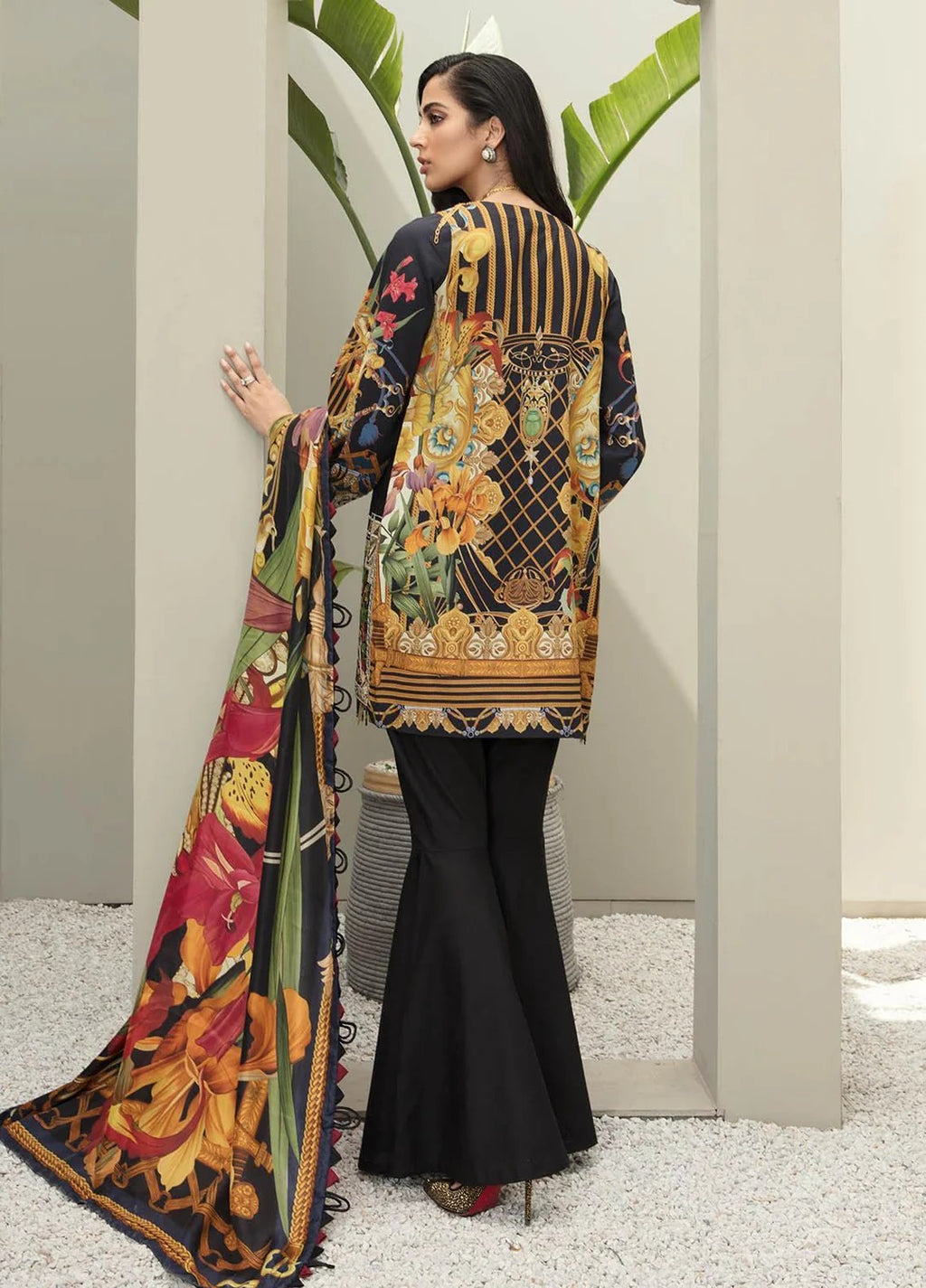 A woman modeling a traditional South Asian outfit, shown from the back. The outfit features a black flared trouser, a long-sleeved tunic with a highly detailed multicolor print in gold, green, and red, and a matching dupatta draped over her shoulder.
