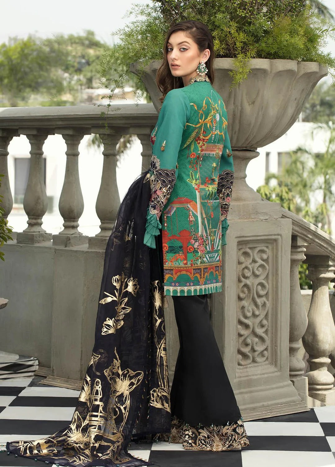 A woman modeling a traditional South Asian outfit with a teal embroidered kurta, black flared pants, and a black dupatta with gold floral patterns, set against an outdoor stone balcony background.