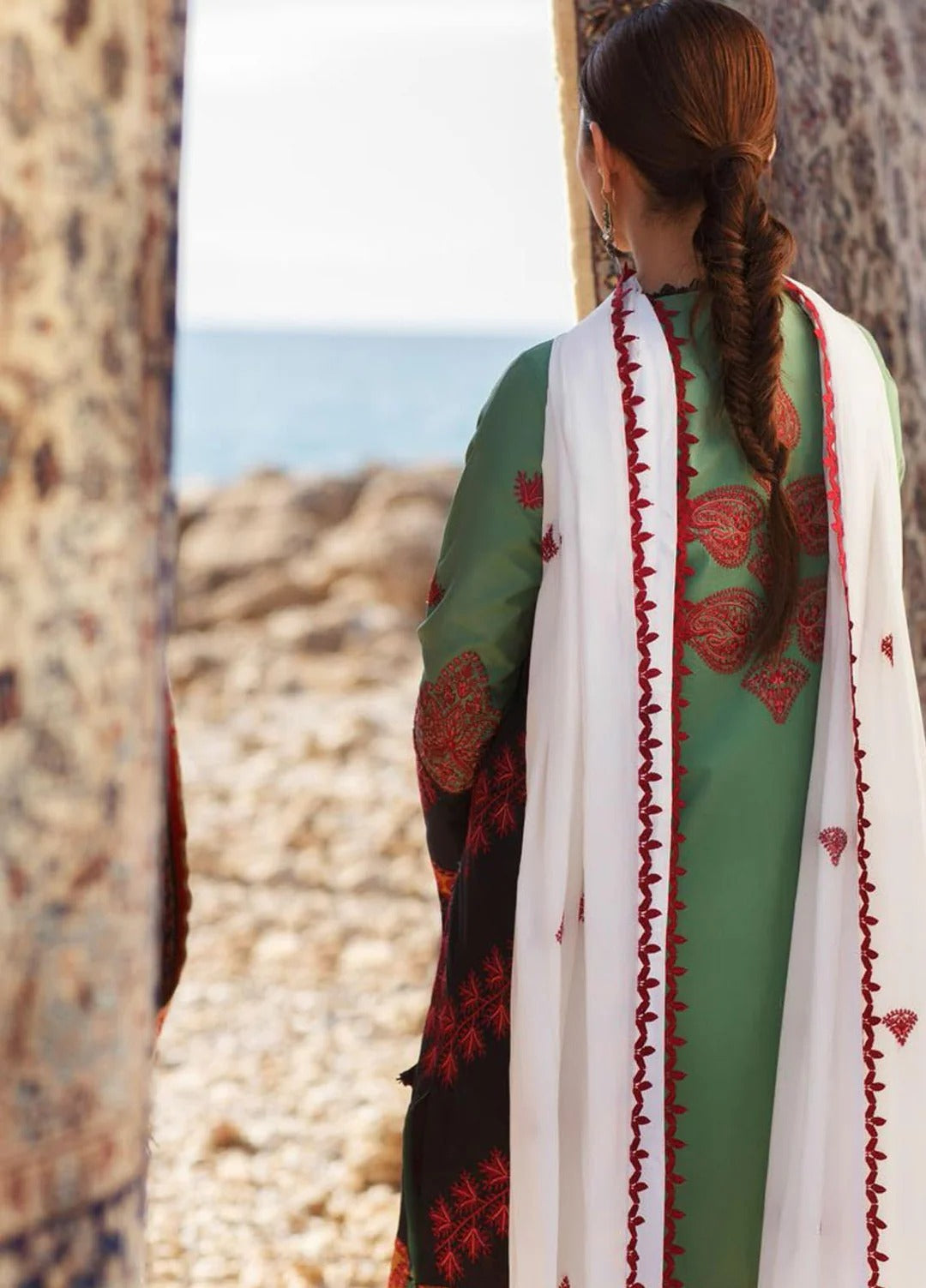 A woman viewed from behind wearing a green traditional outfit with red embroidered patterns, a white dupatta with red border trim, and a black lower panel. She is standing outdoors between textured stone pillars with a rocky beach and sea in the background.
