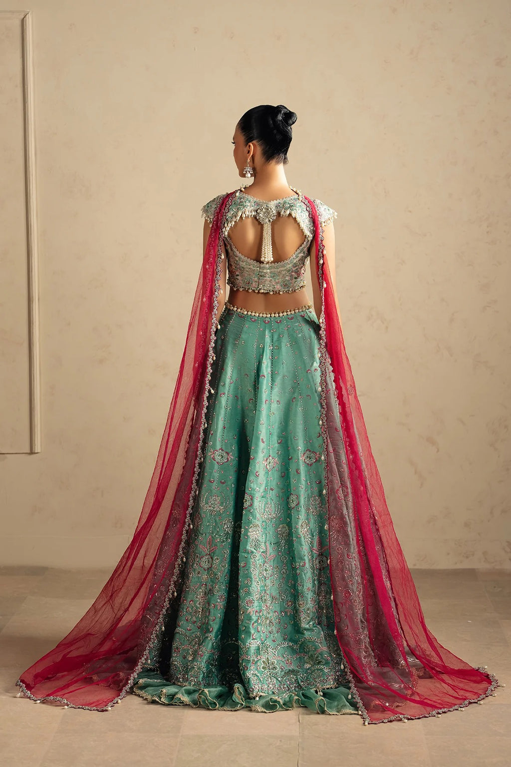 A woman stands with her back to the camera wearing an ornate teal-green embroidered lehenga choli with a long pink dupatta, set against a neutral beige wall.